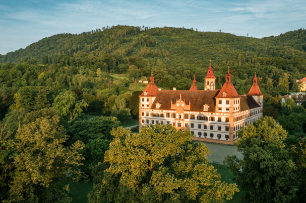 Aerial view of Eggenberg Castle, a large historic building with red roofs surrounded by lush green trees. Located at the foot of a forested hill in Graz, this well-preserved landmark features ornate design elements under clear skies.