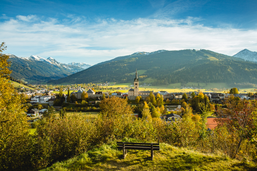 From a wooden bench, you have a picturesque view of the charming town of Radstadt with its striking church tower, surrounded by trees and fields. In the background you can see snow-capped mountains under a blue sky.