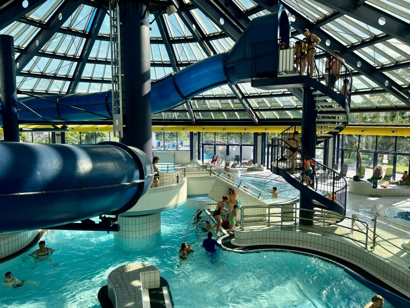 Indoor water park with a large blue water slide that leads into a pool in the Hütteldorfer Bad. People swim, stand in the water, and lounge on lounge chairs. The structure has glass windows and a high, sloped ceiling that lets in natural light. Some people are on the slide staircase.