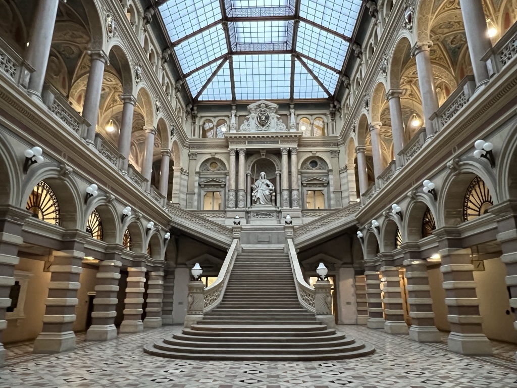 The magnificent interior of the historic Palace of Justice features a central staircase leading up to a large statue surrounded by decorative arches and detailed stonework. The glass ceiling above illuminates the intricate architectural details and patterned floor below, and the Justice Café nearby adds charm.