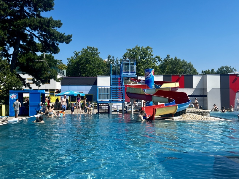 The Ottakringer Bad outdoor pool is teeming with people enjoying the sunny weather. There is a colorful water slide with several twists and turns. Some people use the slide while others relax in the water. Green trees and a clear blue sky create the perfect backdrop.