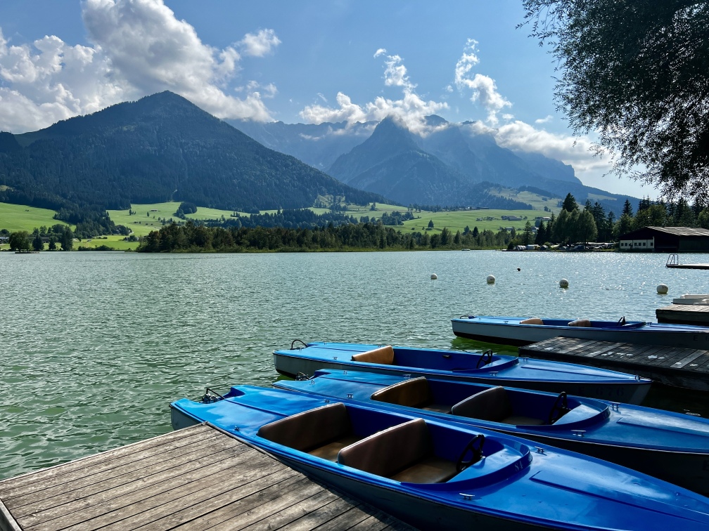 Blaue Ruderboote liegen am Rande eines ruhigen Sees mit malerischen Bergen und üppigem Grün im Hintergrund und zeigen die Naturidylle des Kaiserwinkels Tirol. Der Himmel ist teilweise bewölkt, was die ruhige und malerische Atmosphäre von Walchsee noch verstärkt.