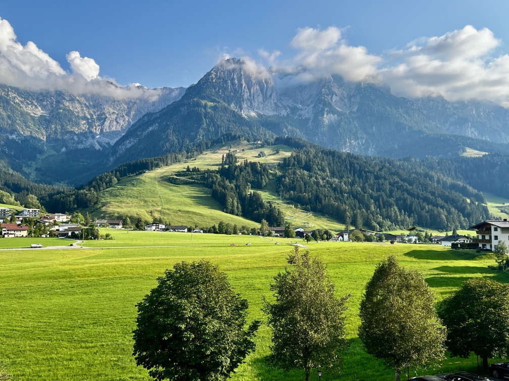 A picturesque landscape with lush green fields, scattered trees and a small town in the foreground. In the background, the Zahme Kaiser Mountains, partially covered in clouds, tower over the scenery, with a clear blue sky above. A mountain railway winds its way up the slopes, lending a special charm to this tranquil setting.