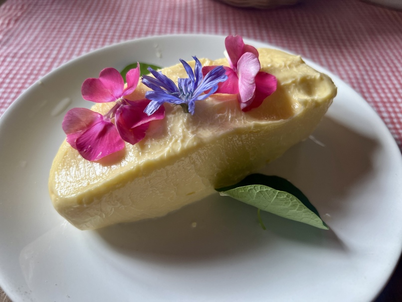 A smooth, yellow pat of butter on a white plate is garnished with pink and purple flowers reminiscent of grandma's times and a few green leaves. The plate sits on a red and white checked tablecloth, evoking memories of butter making in simpler times.