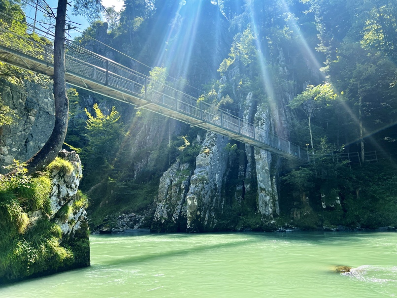 A metal suspension bridge crosses the turquoise Tiroler Ache river between cliffs in a forested area. Sunlight filters through the trees, creating shafts of light that illuminate the tranquil scene. Moss-covered rocks can be seen in the foreground, hinting at possible adventures such as rafting nearby.