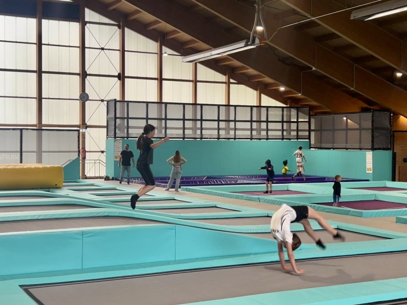 People are having fun in a large indoor trampoline park. In the foreground, a person is doing a somersault, while in the background others are jumping and playing. The facility, which is reminiscent of the hustle and bustle at the Zahmer Kaiser in Walchsee, has several trampolines with safety pads.