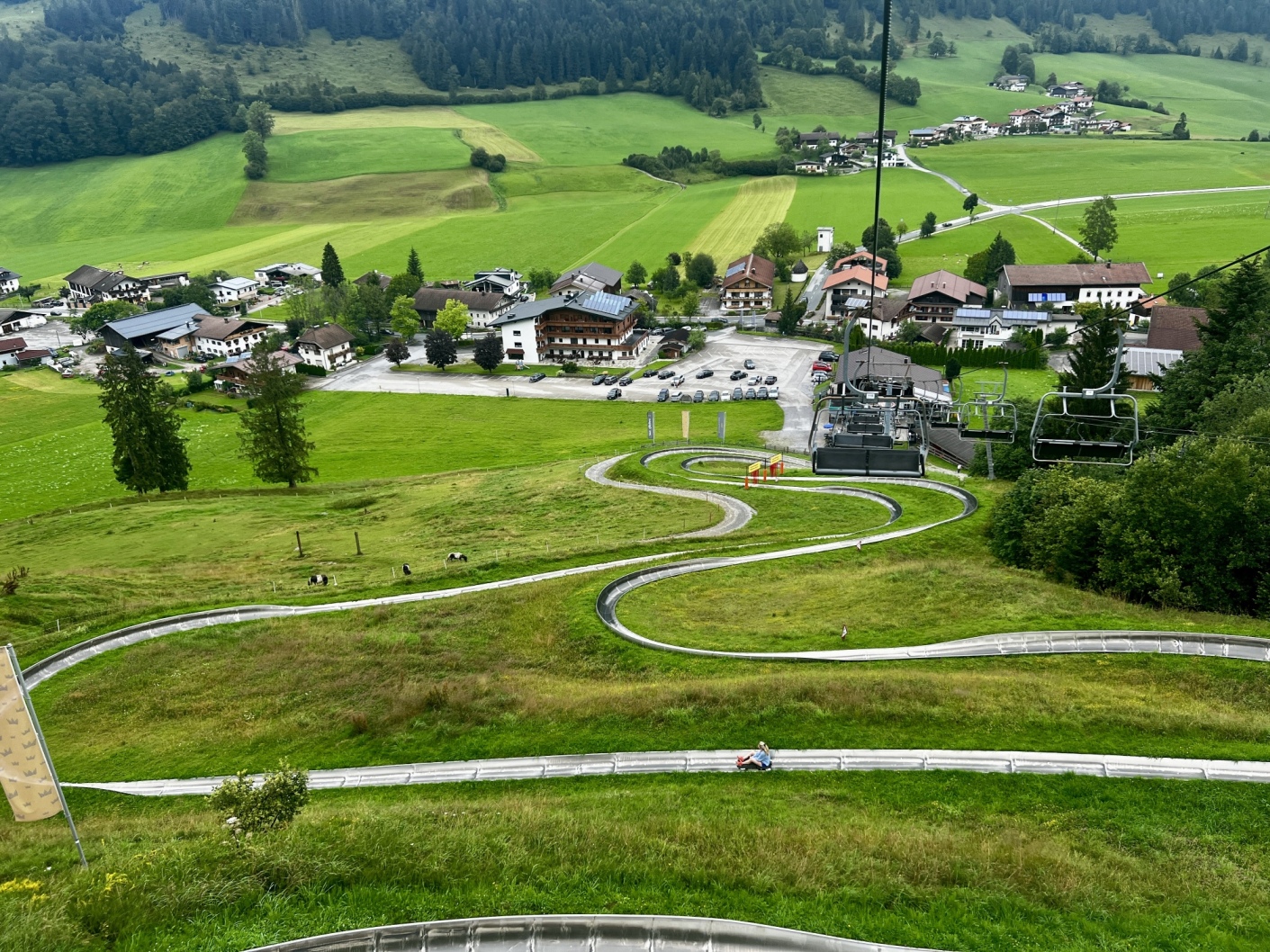A winding toboggan run leads down a grassy hill, with the adventure mountain railway chairlift running alongside it. Below lies a small village with various buildings and parked cars in a green valley, surrounded by trees and hills, against the majestic backdrop of the Zahmer Kaiser.