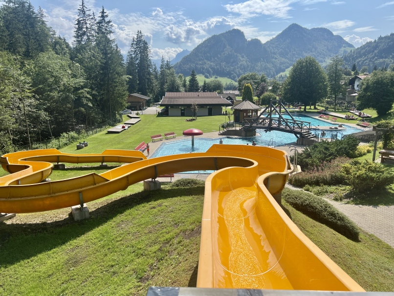 An outdoor water park in Kössen features a yellow slide with twists and turns. The adventure forest swimming pool park includes several pools, lounge areas and lawns surrounded by trees and mountains in the background under a partly cloudy sky. Visitors can be seen enjoying the amenities.