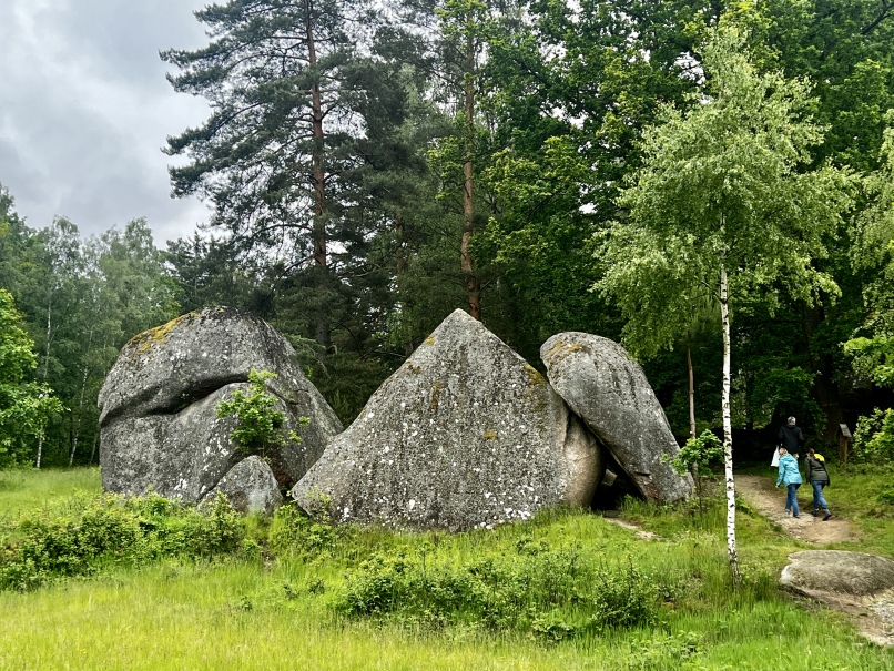 Zwei Menschen gehen auf große, moosbedeckte Felsbrocken zu, die als Wackelsteine bekannt sind, inmitten eines üppigen grünen Waldes im Naturpark Blockheide. Der Himmel scheint bedeckt zu sein. Bäume umgeben das Gebiet und tragen zur dichten Waldatmosphäre bei. Der Weg ist schmal, mit Gras und kleinen Büschen auf beiden Seiten, was die Kulisse für ein Abenteuer schafft.