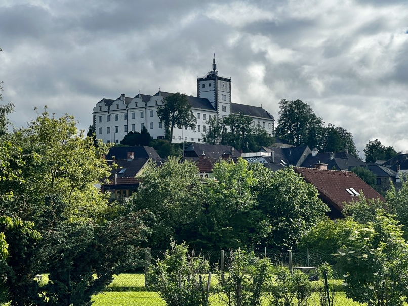 A white multi-story building with a distinctive central tower stands on a hill surrounded by lush green trees and smaller buildings in the foreground. The sky is covered with clouds, providing a dramatic backdrop to the historic splendor of Weitra Castle in the picturesque Waldviertel region.