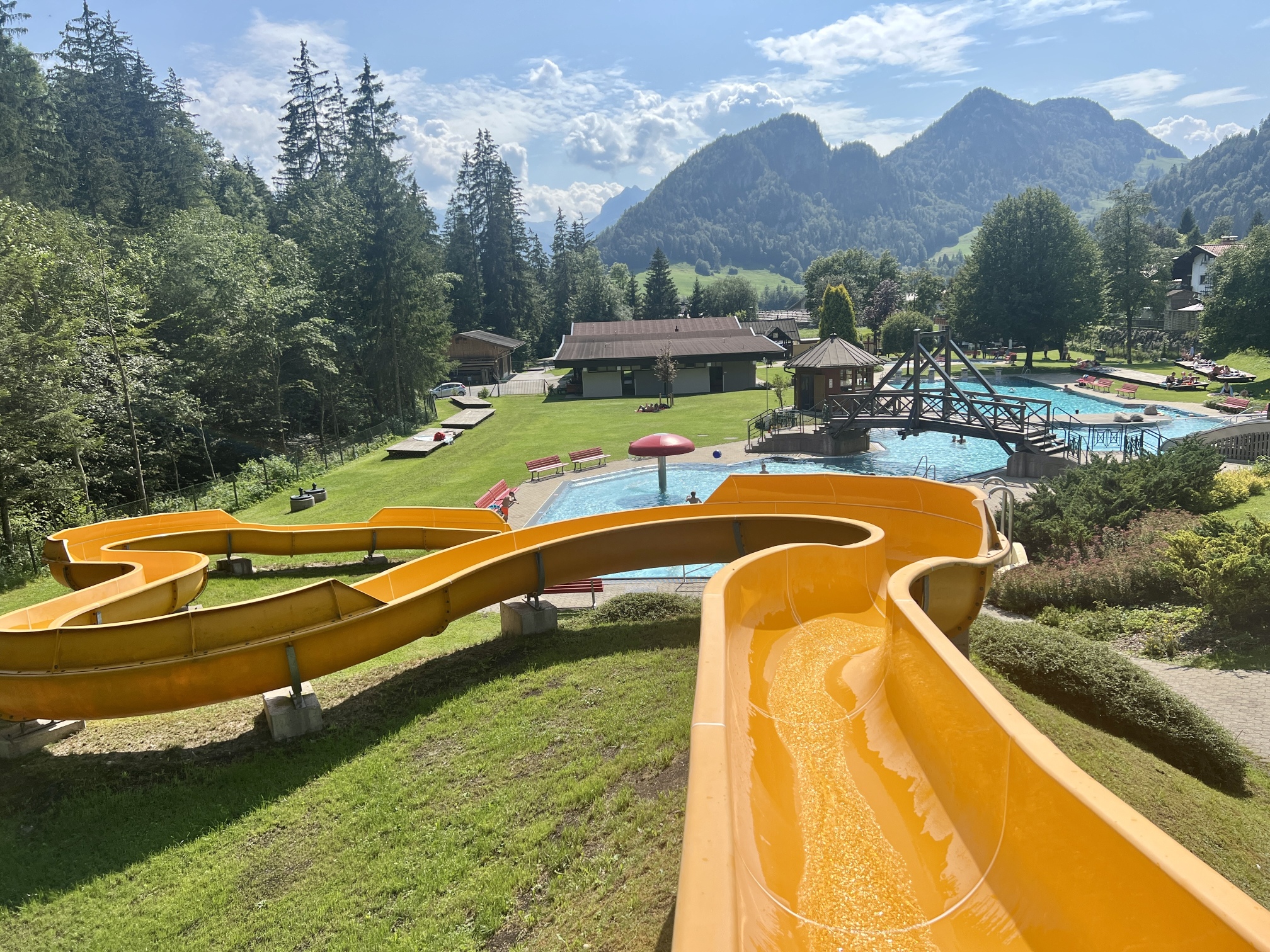 Ein malerischer Wasserpark im Freien, perfekt für Ausflüge mit Kindern, verfügt über eine große orangefarbene Wasserrutsche. Der Park Familienparadies Kaiserwinkl umfasst einen Swimmingpool, Sitzbereiche mit grünem Gras, Bäumen und einer Berglandschaft im Hintergrund. Über uns sind klarer blauer Himmel und vereinzelte Wolken zu sehen.