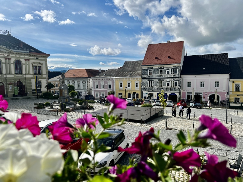 A cobblestone town square is surrounded by colorful buildings. Pastel-colored facades in shades of yellow, pink, blue and white add charm to Weitra, Austria's oldest brewing town. In the foreground, bright pink and white flowers bloom under a partly cloudy sky.