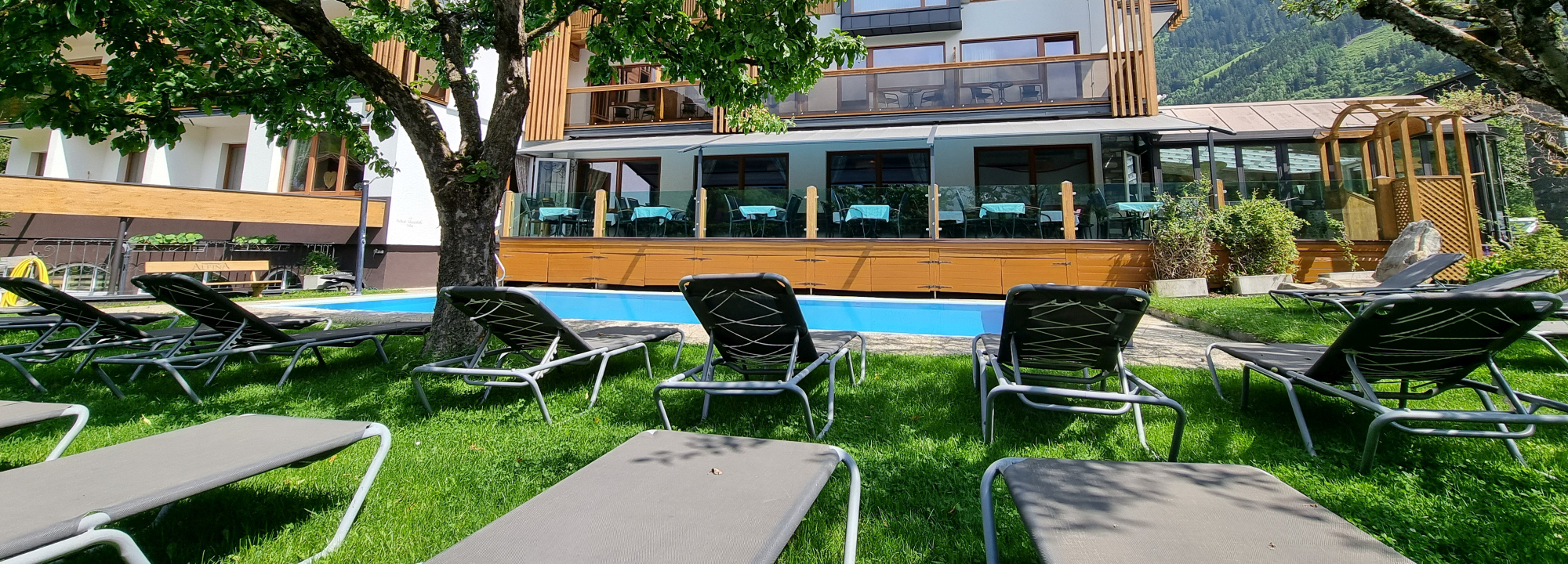 In front of a rectangular pool at Hotel Alpina Rauris there is a lawn with several sun loungers. The building behind the pool has a glass-enclosed dining area with blue tablecloths and offers partial views of an adjacent terrace and the nearby mountains.