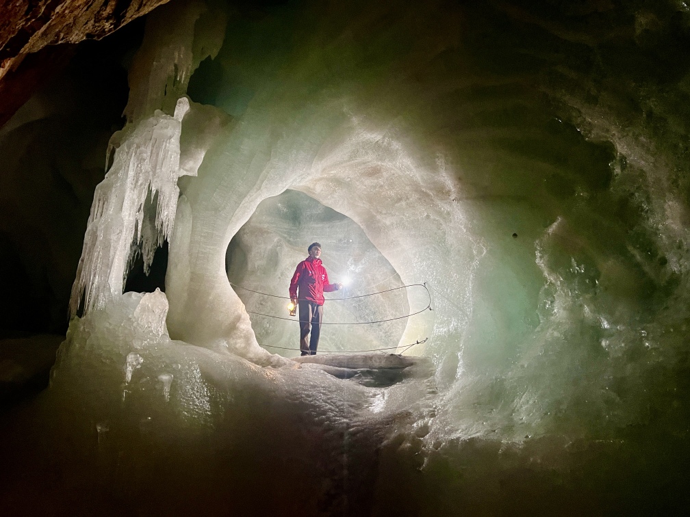 A person in a red jacket stands on a metal railing bridge in the Eisriesenwelt near Werfen. The cave walls are covered with thick ice formations and icicles, and the person is holding a flashlight that illuminates the surroundings.