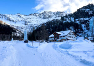 Eine verschneite Berglandschaft mit einer großen Holzhütte, umgeben von schneebedeckten Bäumen. Der Himmel ist teilweise bewölkt und eisige Formationen sind sichtbar. Schneebedeckte Berge ragen deutlich im Hintergrund hervor und schaffen eine ruhige Winterlandschaft.