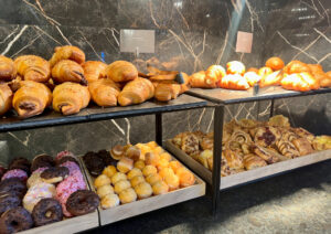 Eine Bäckerei-Auslage mit verschiedenen Backwaren: In den oberen Regalen befinden sich Croissants, in den unteren Regalen verschiedene Donuts und Backwaren. Der Hintergrund ist eine dunkle Marmorwand.