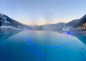 Panoramablick auf einen Infinity-Pool, in dem sich der Himmel spiegelt, mit schneebedeckten Bergen im Hintergrund. Die untergehende Sonne wirft ein sanftes Licht und rechts ist ein modernes Gebäude mit großen Fenstern zu sehen. Die Atmosphäre ist heiter und ruhig.