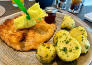 Ein Teller Schnitzel mit einer dekorativ eingewickelten Zitrone darauf, serviert mit gewürzten Salzkartoffeln. Neben dem Schnitzel steht ein kleines Glas mit roter Marmelade. Im Hintergrund sind Getränke auf einem Tisch zu sehen.