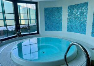 Innen-Whirlpool mit klarem Wasser, vor einer Wand mit blauen Mosaikfliesen. Große Fenster mit Milchglasplatten geben teilweise den Blick auf eine verschneite Außenlandschaft frei. Auf der rechten Seite der Wanne befindet sich ein Metallhandlauf.